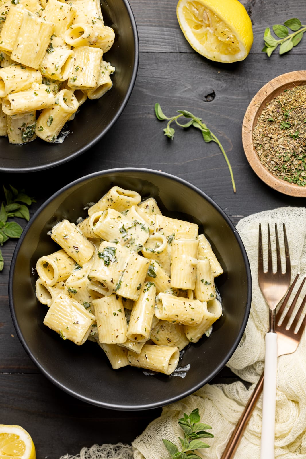 Two bowls of pasta on a black table with forks, lemons, and herbs.