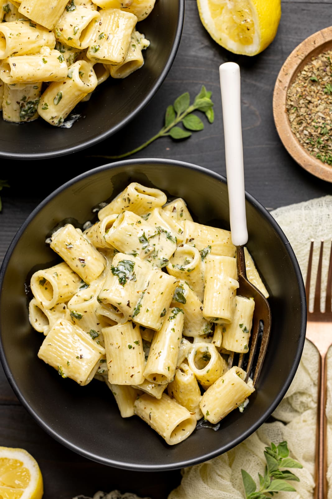 Up close shot of pasta in a black bowl with a fork, lemons, and herbs + seasonings. 