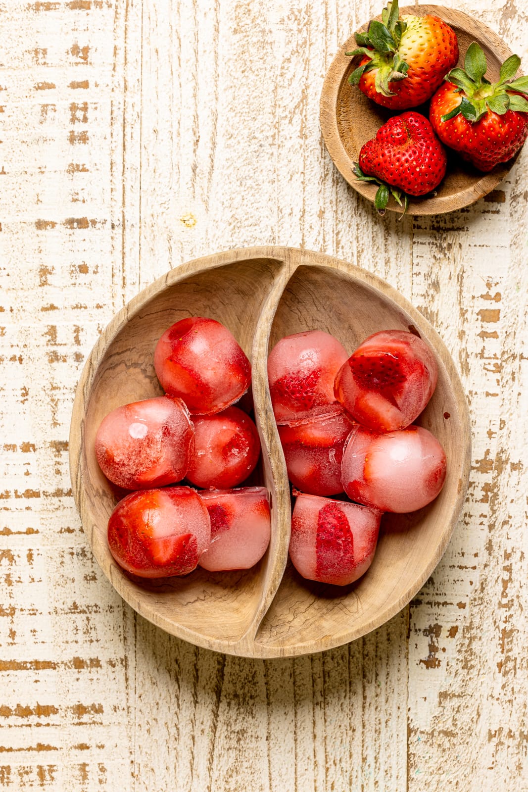 Strawberry ice cubes in a wood bowl on a white table with fresh strawberries.