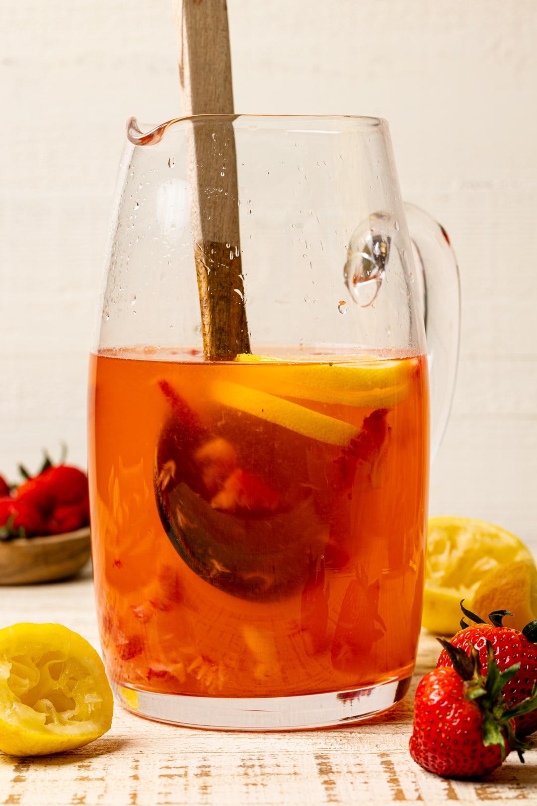 Strawberry lemonade in a large glass pitcher with a wooden spoon on a white table with strawberries and lemons.