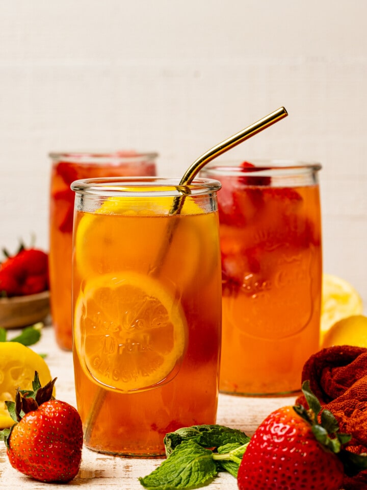 Three tall glasses of lemonade with a straw on a white wood table with strawberries and lemons.