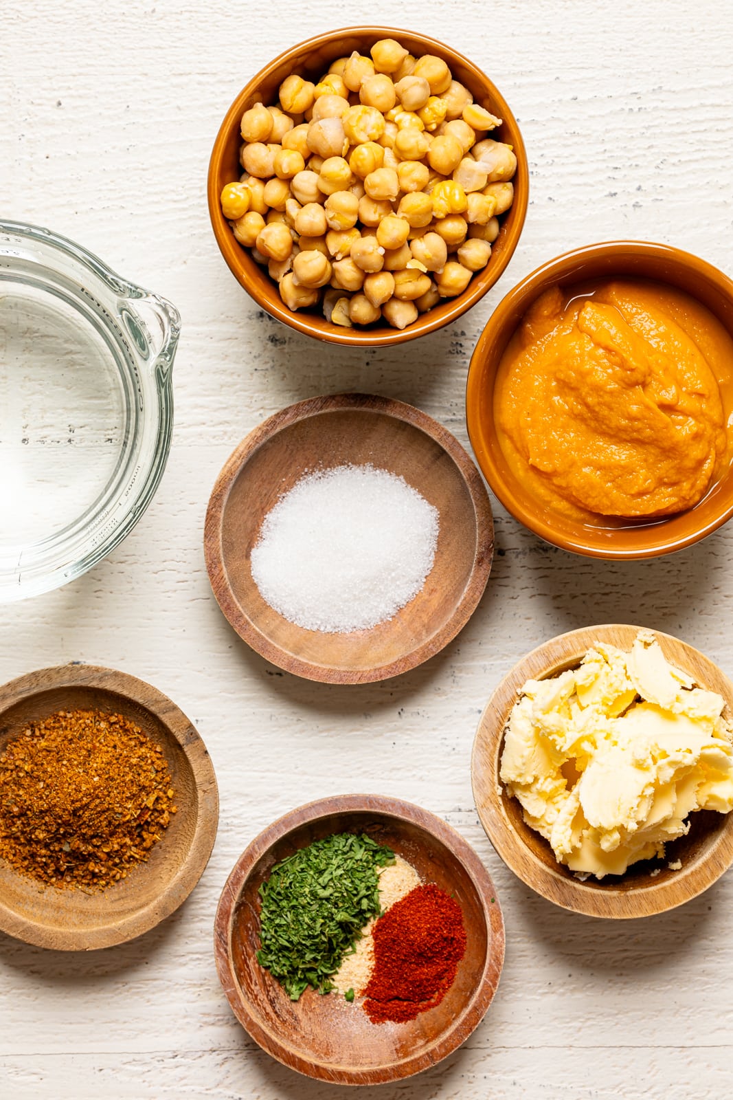 Ingredients on a white wood table including chickpeas, sweet potato puree, water, herbs and seasonings, butter, sugar, and cajun seasoning.