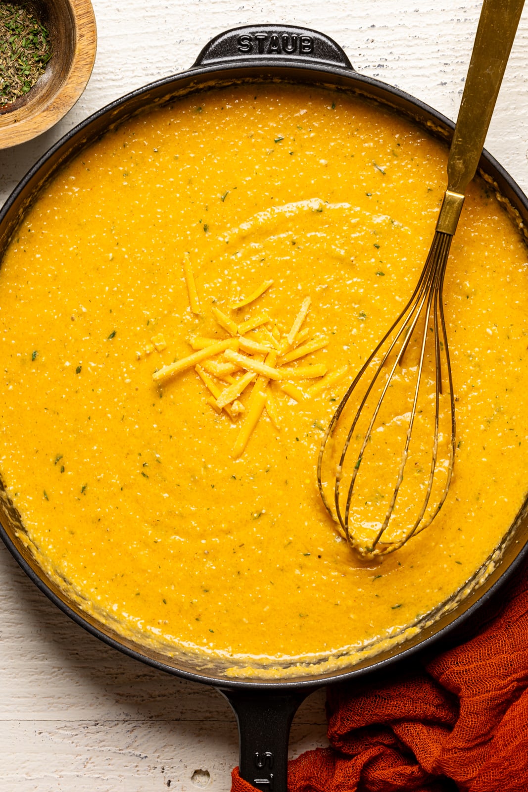 Creamy grits in a black skillet with a gold whisk on a white wood table with a red napkin.