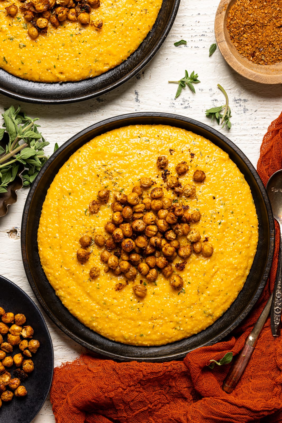 Chickpeas and grits in two black plates with a side of chickpeas and herbs on a white table.