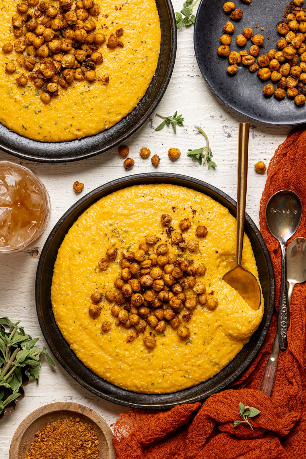 Spoon scooping chickpeas and grits from two black plates with a side of juice and chickpeas on a white wood table.