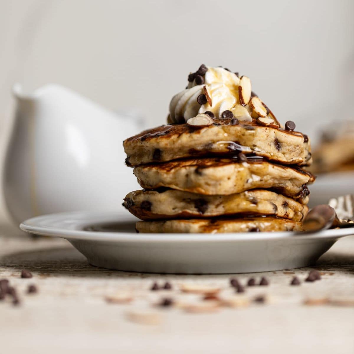 Stack of pancakes in a white plate with chocolate chips + almonds on a white wood table.
