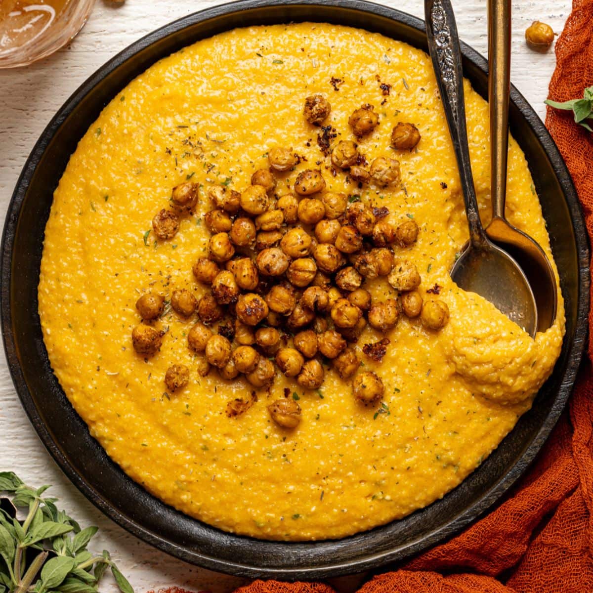 Chickpeas and grits on a black plate with two spoons and a red napkin and herbs.