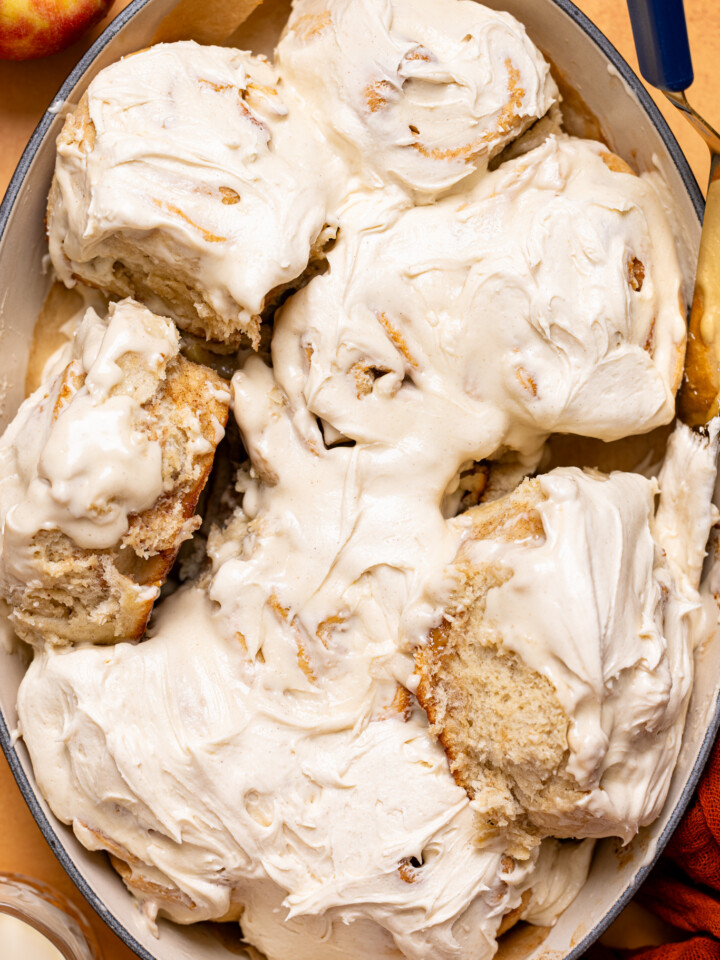 Frosted apple pie cinnamon rolls in a baking dish with a spatula, milk, and apple.