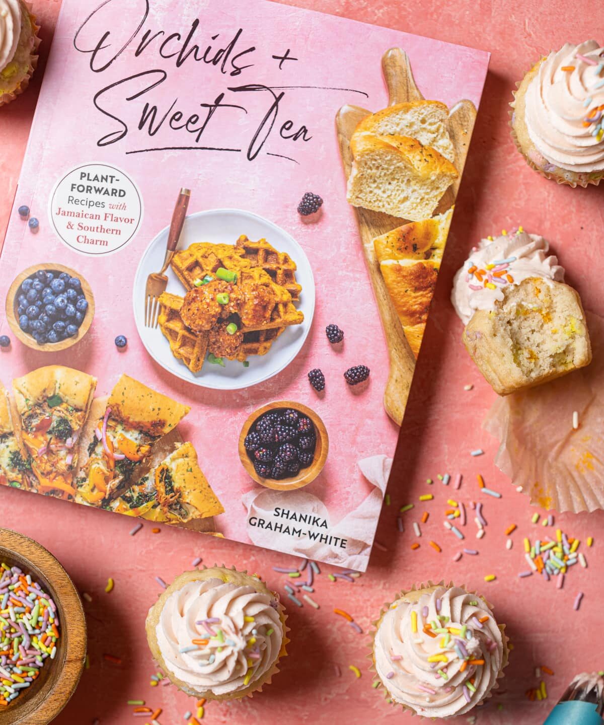 Cupcakes, sprinkles, and the Orchid + Sweet Tea cookbook on a pink table