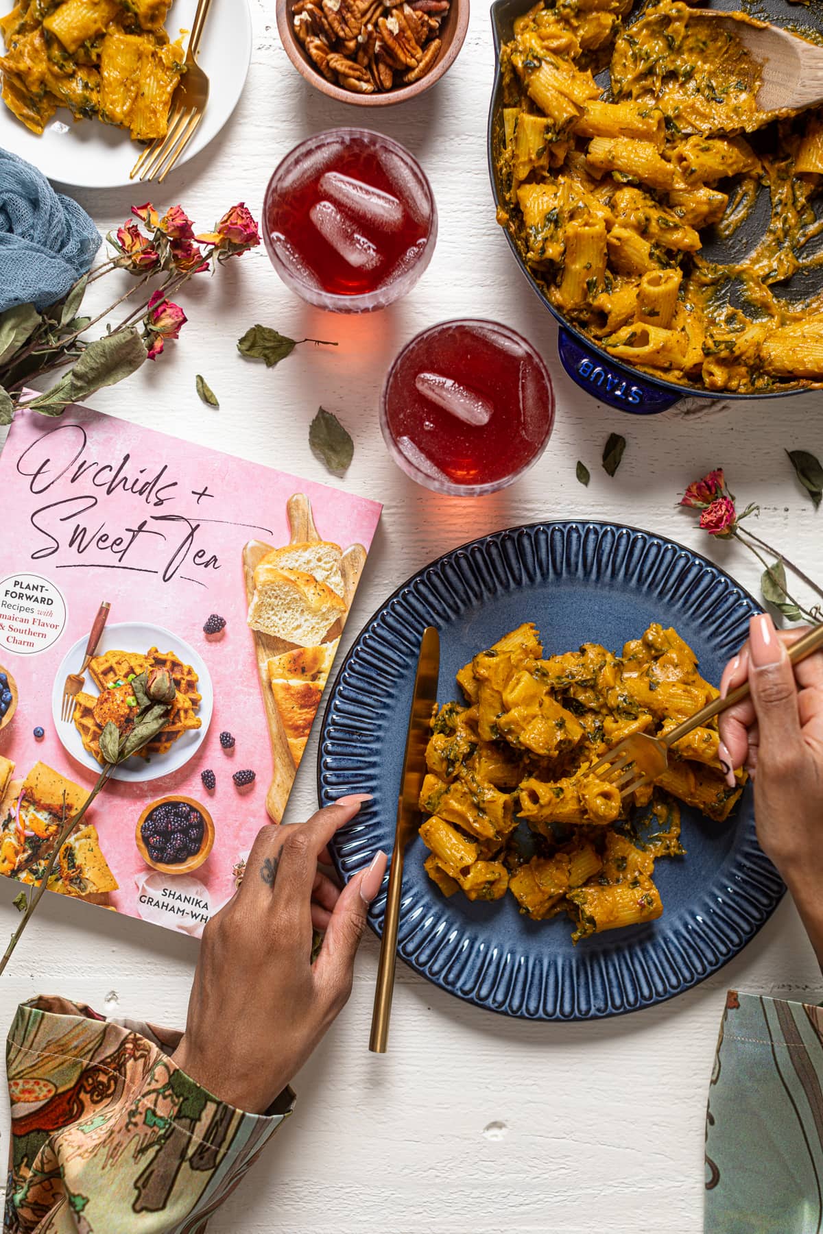 Blue plate and skillet of pasta on a table with red drinks and the \"Orchids + Sweet Tea\" book