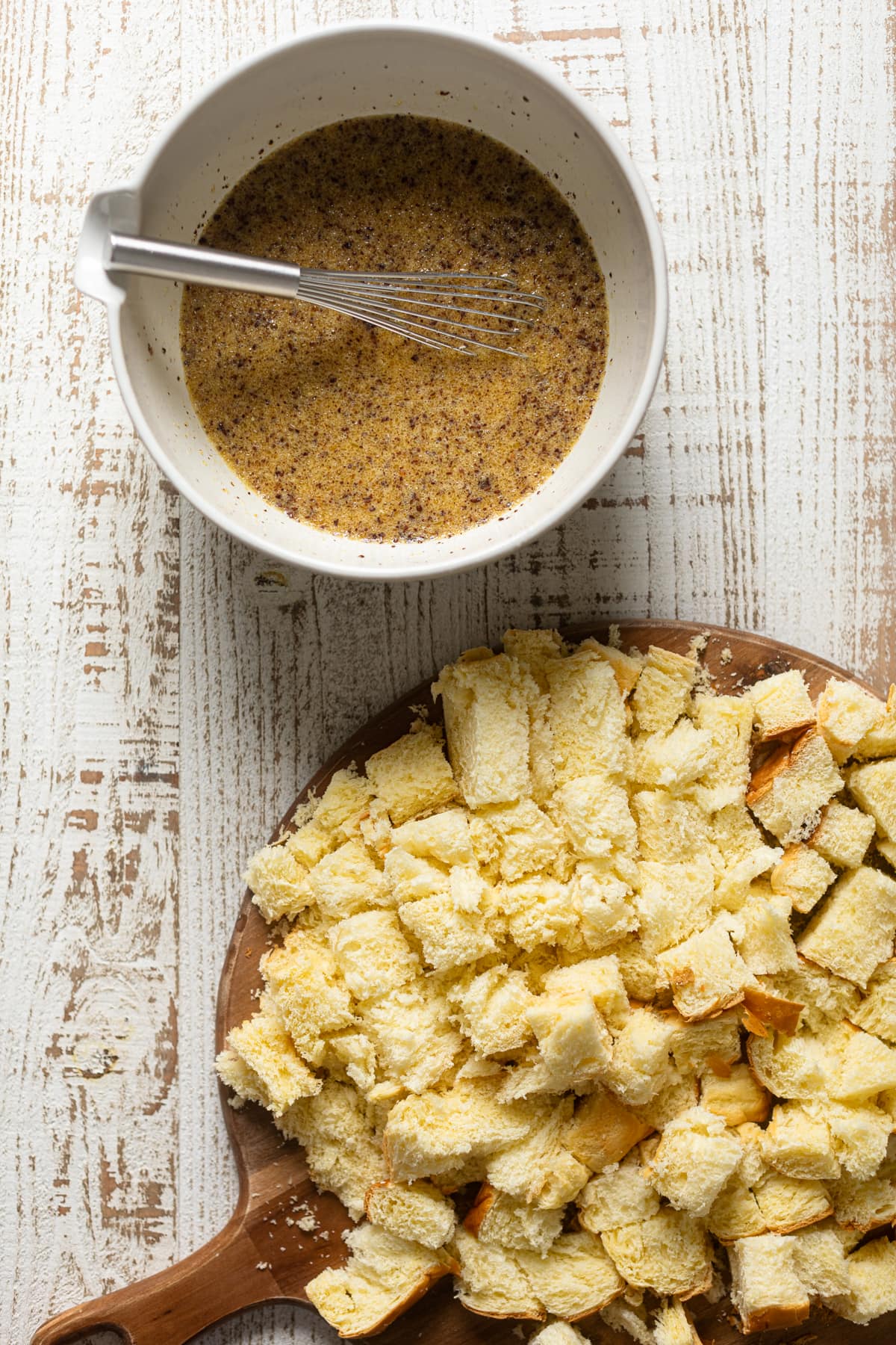 Bowl of egg mixture next to a wooden board topped with chopped bread