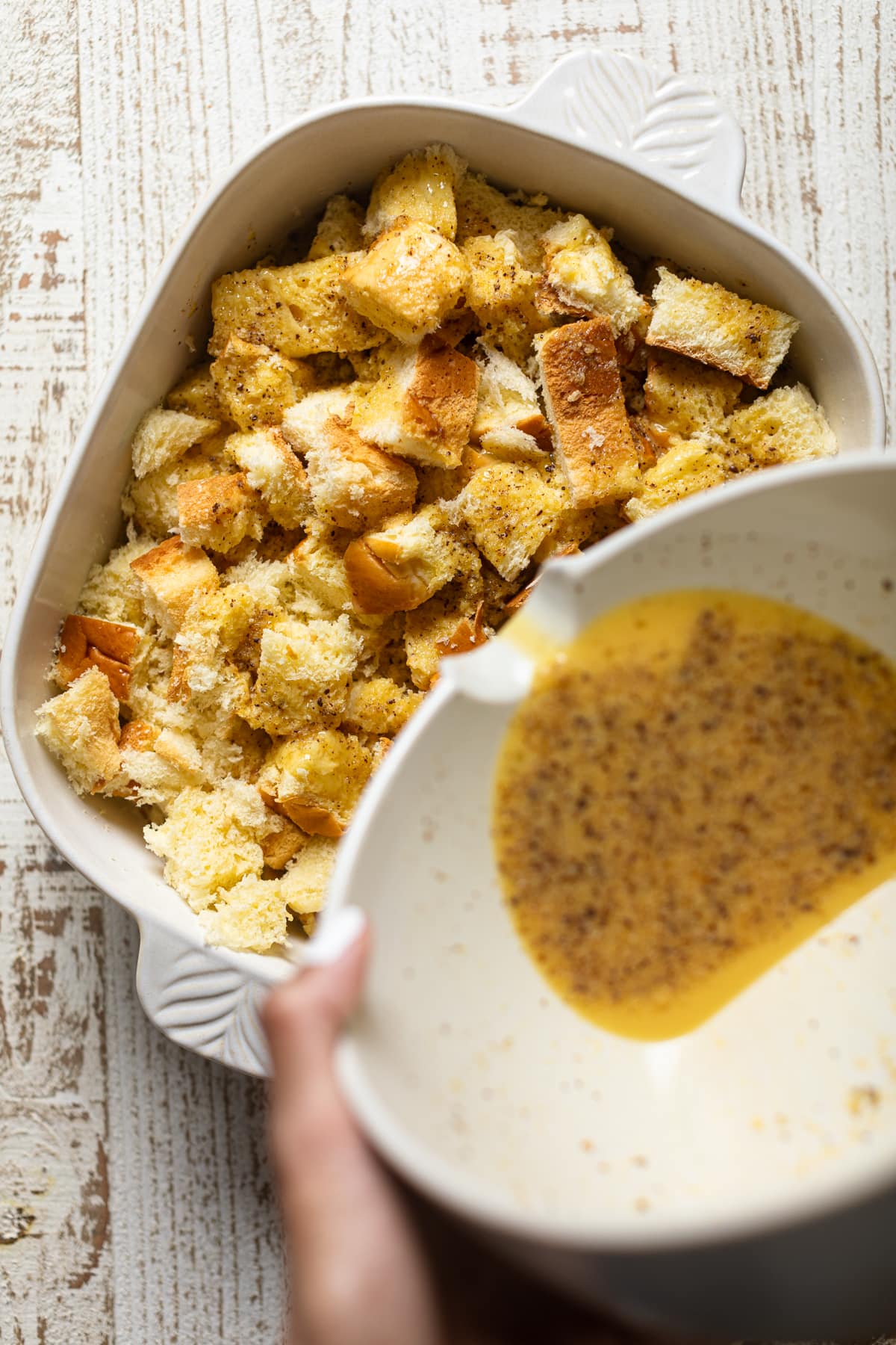 Bowl pouring egg mixture onto a baking pan of bread pieces