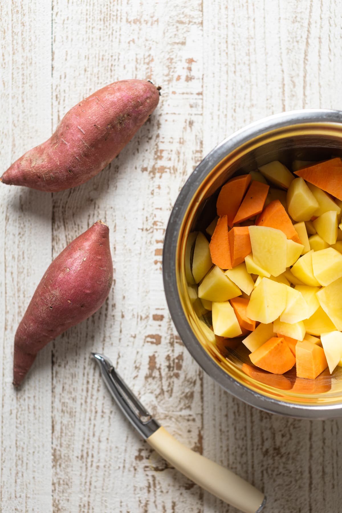 Bowl of chopped and peeled potatoes and sweet potatoes next to sweet potatoes and a potato peeler