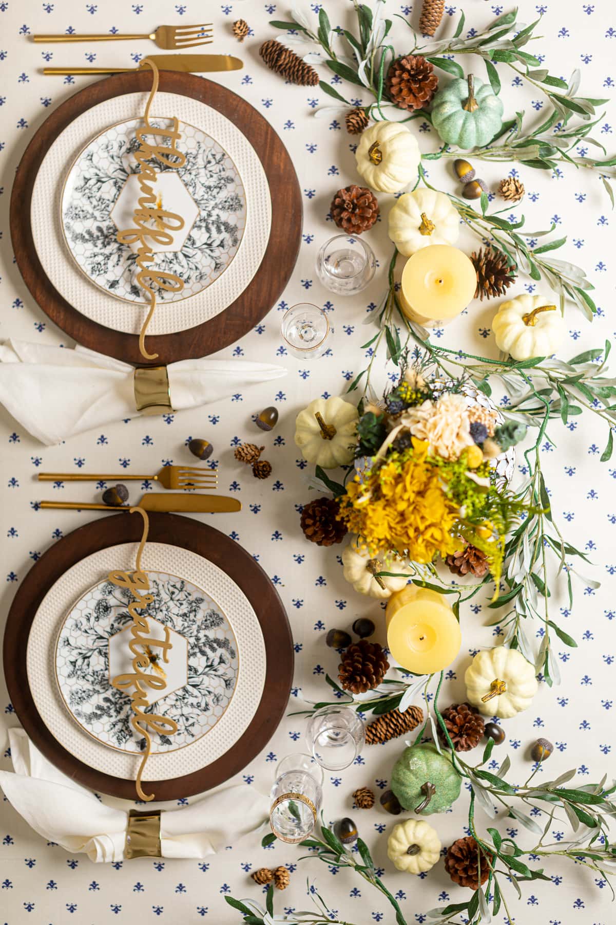 Decorative place settings, pumpkins, candles, flowers, and pine cones near plates of carrots and rolls on a mostly-white tablecloth