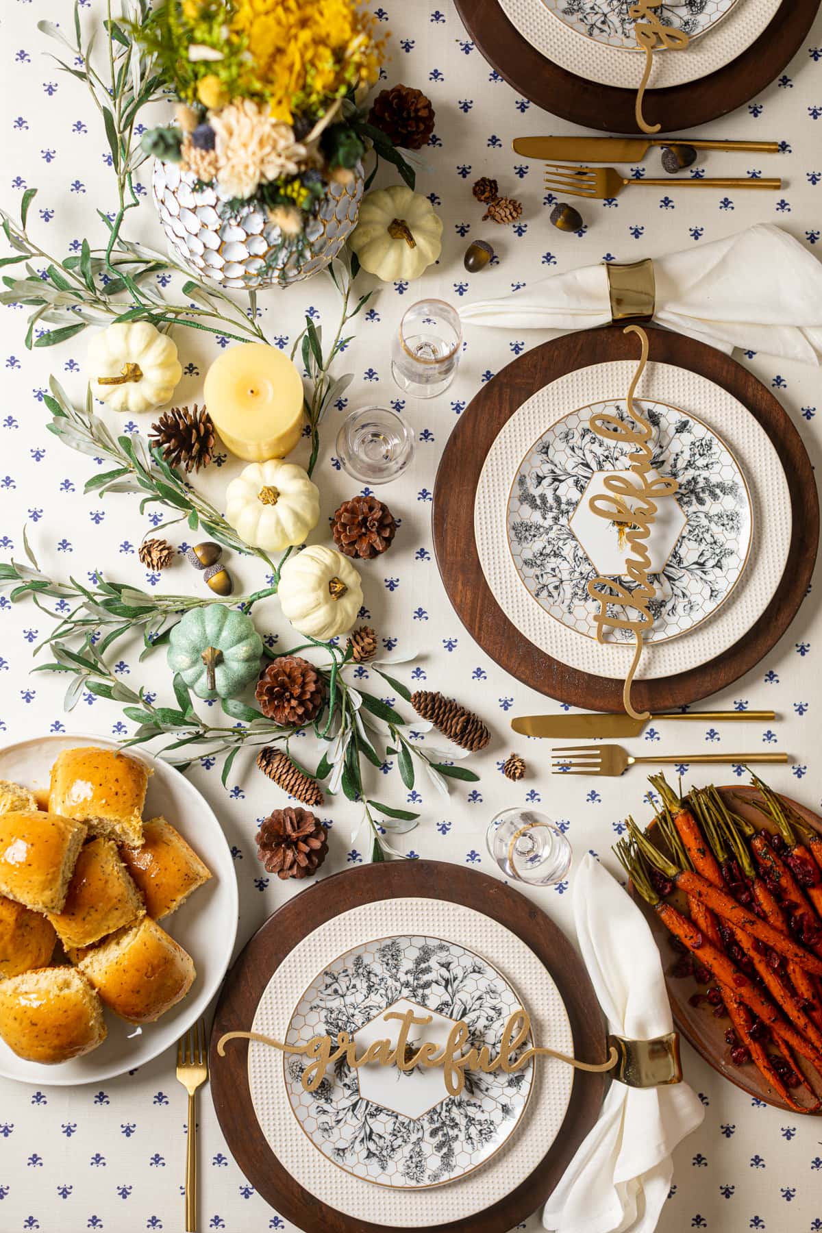 Decorative place settings, pumpkins, candles, and pine cones near plates of carrots and rolls on a mostly-white tablecloth