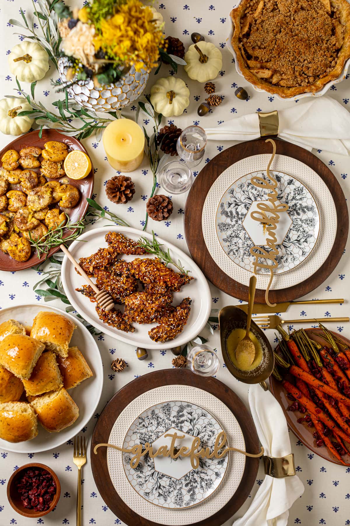 Decorative place settings, pumpkins, candles, and pine cones near plates of beautiful food on a mostly-white tablecloth