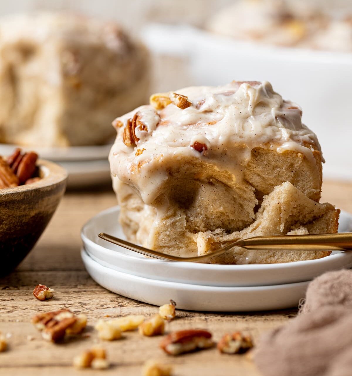 Up close shot of cinnamon roll on a stack of white plates with a gold fork and cinnamon rolls in the background.