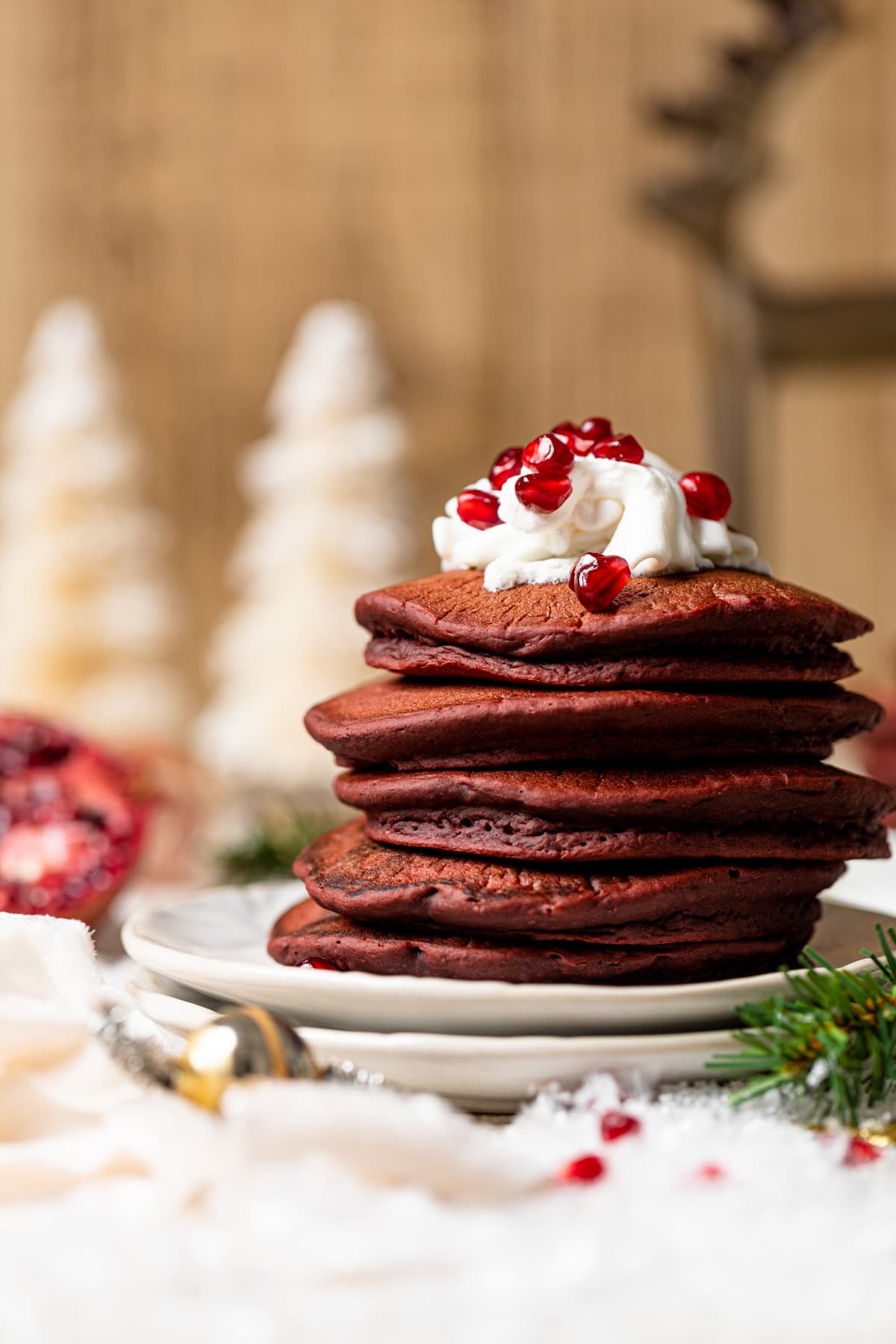 Stack of Fluffy Red Velvet Pancakes topped with whipped cream and pomegranate seeds on two plates near Christmas decorations