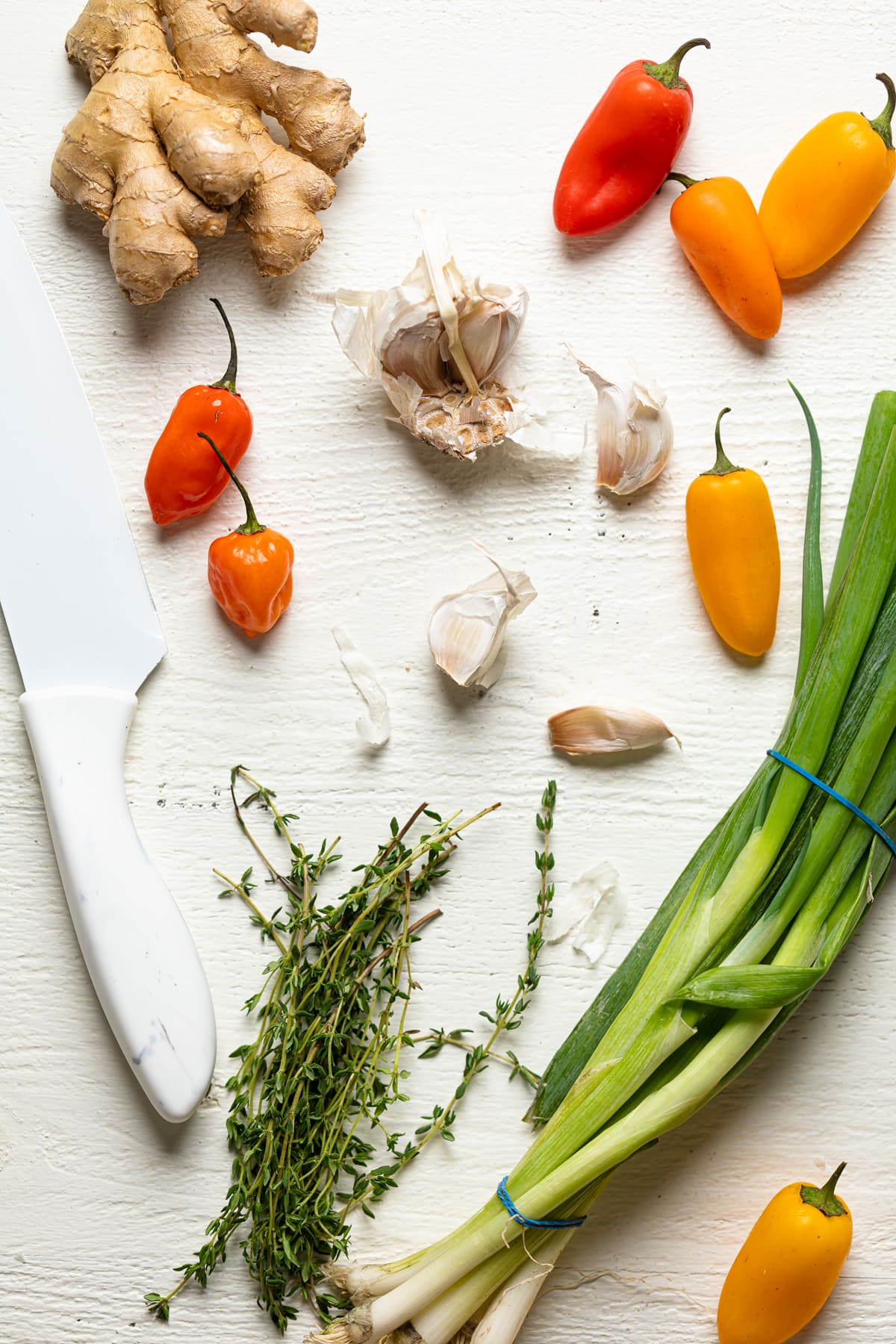 Thyme, green onions, garlic, scotch bonnet peppers, ginger, and a knife on a white surface