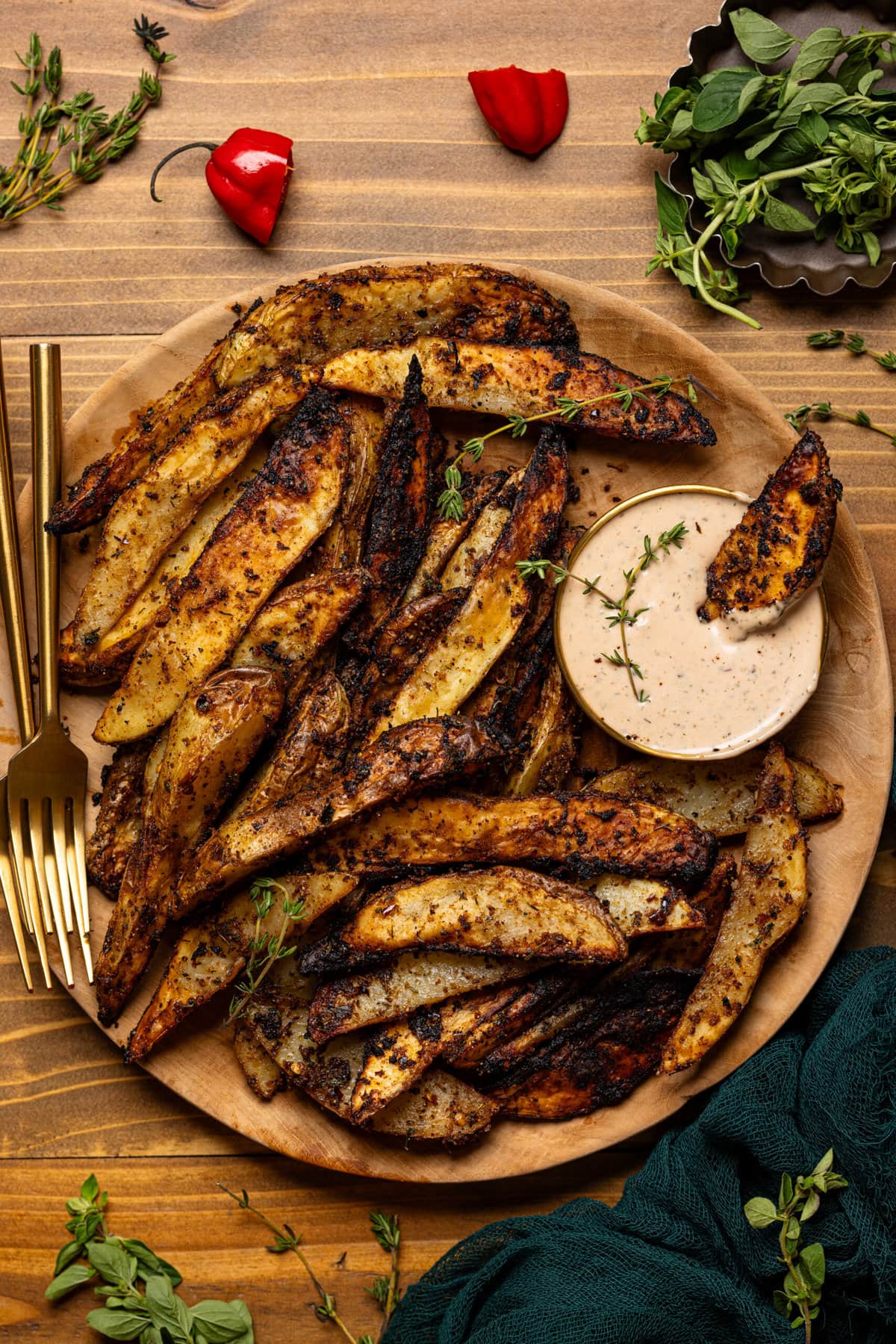 Jerk wedges on a brown wooden plate with a dipping sauce, fork, and herbs.