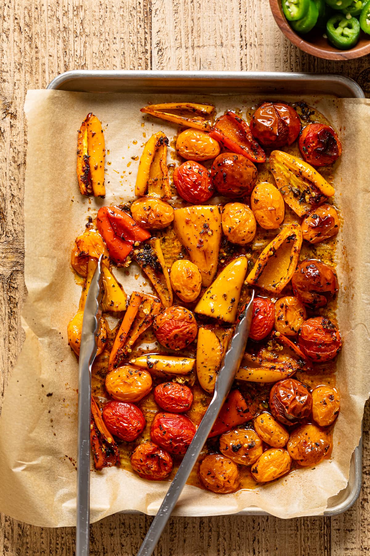 Tongs on a baking sheet of roasted peppers and tomatoes