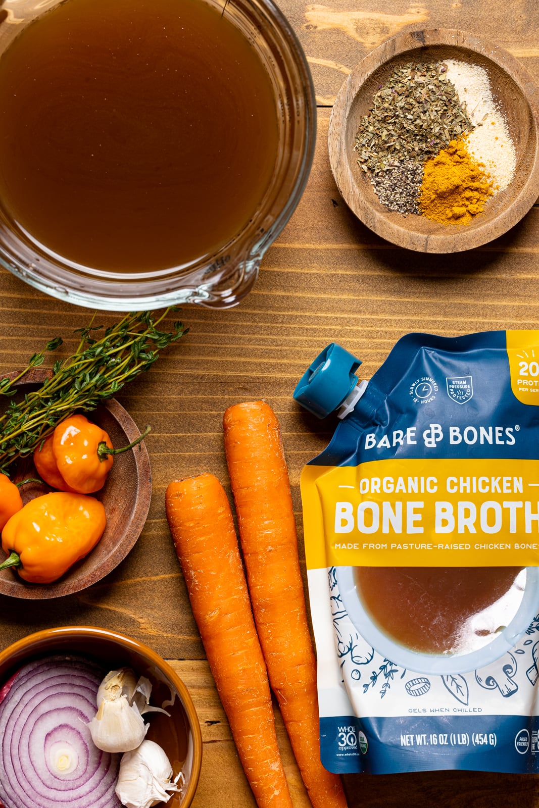 Ingredients on a brown wood table including chicken stock, bone broth, carrots, onions, thyme, scotch bonnet, and seasonings and herbs.