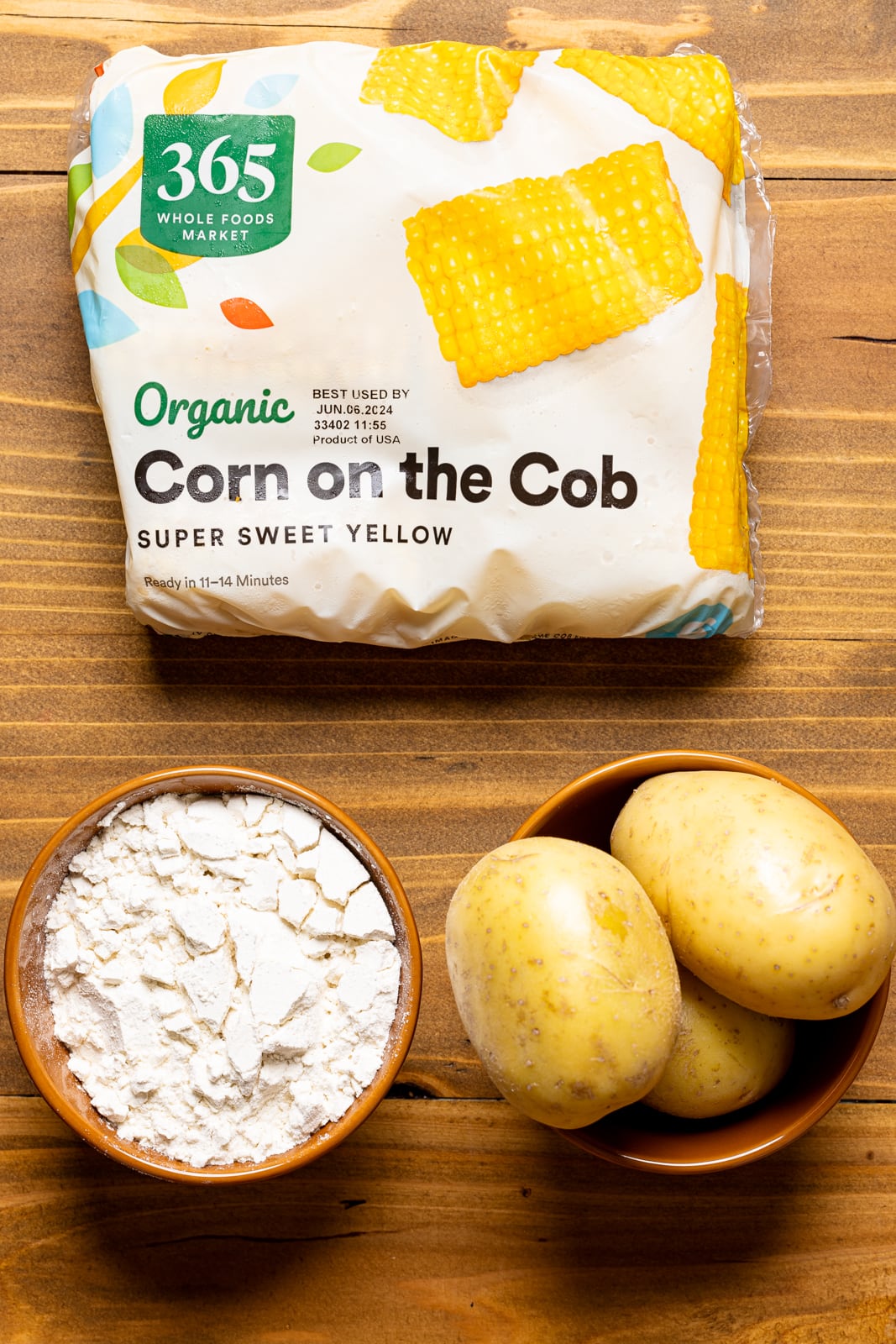 Ingredients on a brown wood table including corn, potatoes, and flour.