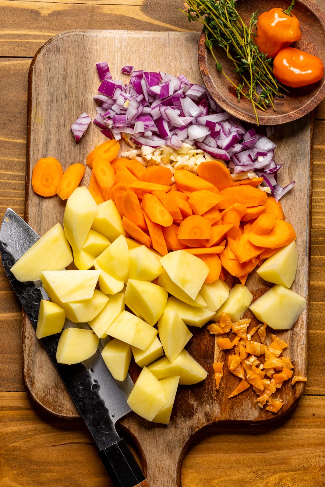 Ingredients on a brown wood cutting board including chopped carrots, potatoes, onions, scotch bonnet, and thyme.