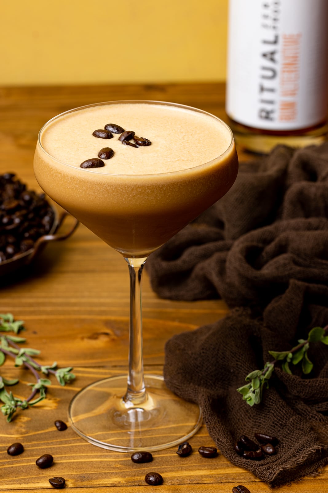 Espresso martini in a glass on a brown wood table with a brown napkin, coffee beans, and Rum alternative. 