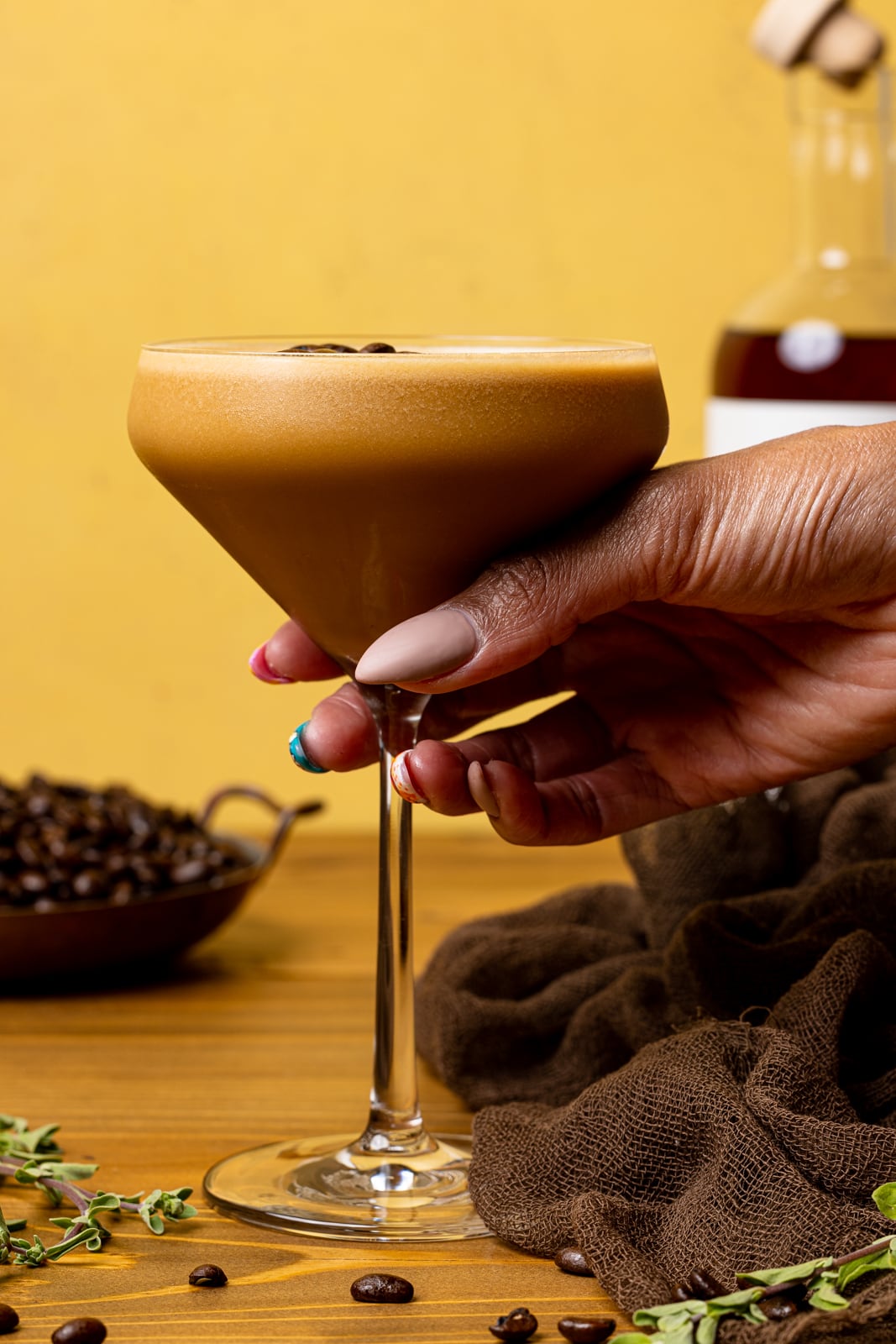 Espresso martini in a glass on a brown wooden table with a yellow background being held. 