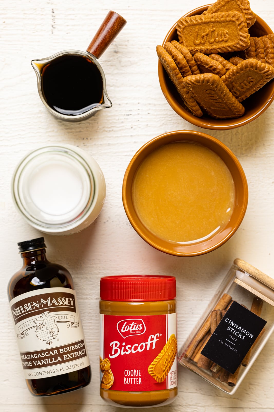 Ingredients on a white table including brewed coffee, biscoff cookies, milk, vanilla, biscoff cookie butter, caramel sauce, and cinnamon.