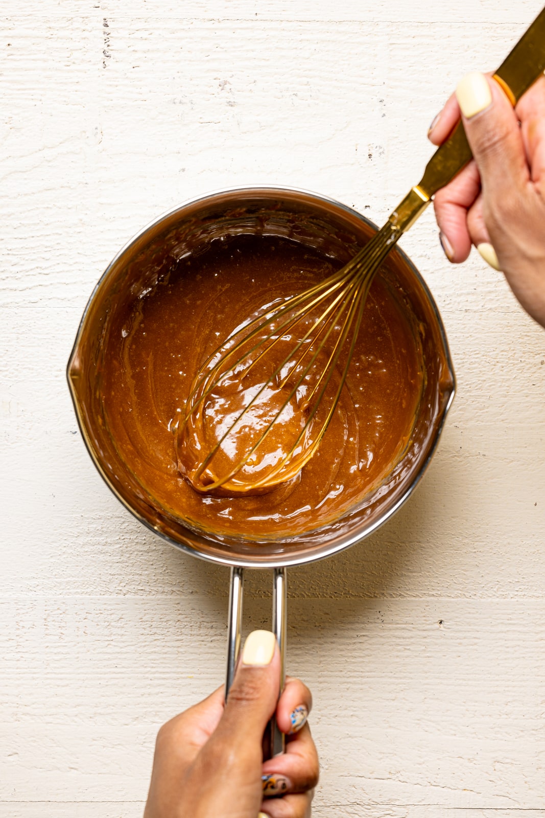 Melted cookie butter in a silver saucepan with a whisk on a white table.