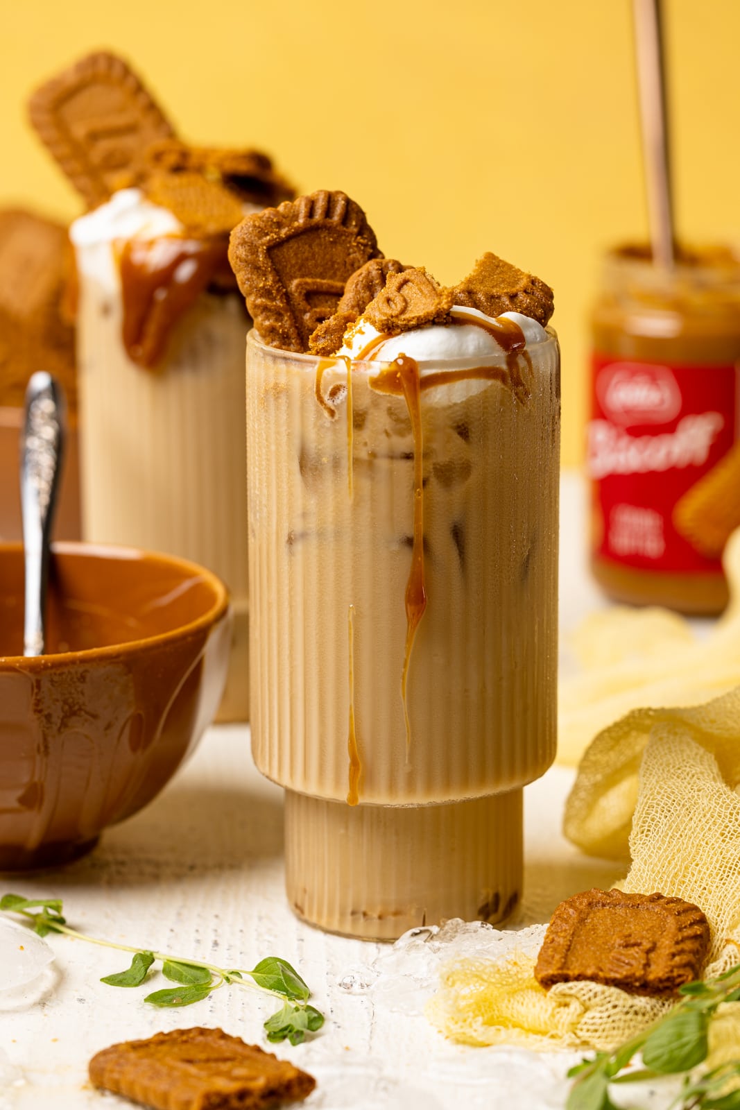 Two glasses with lattes, biscoff cookies, and cookie butter in the background on a white table and yellow background. 