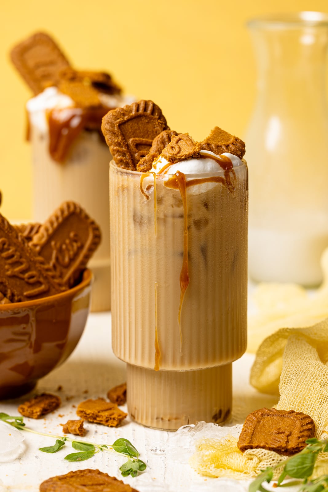 Two glasses with lattes, biscoff cookies, and cookie butter in the background on a white table and yellow background.