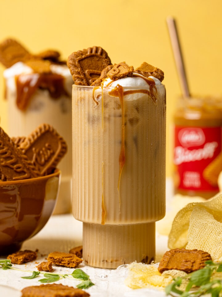 Two glasses with lattes, biscoff cookies, and cookie butter in the background on a white table and yellow background.