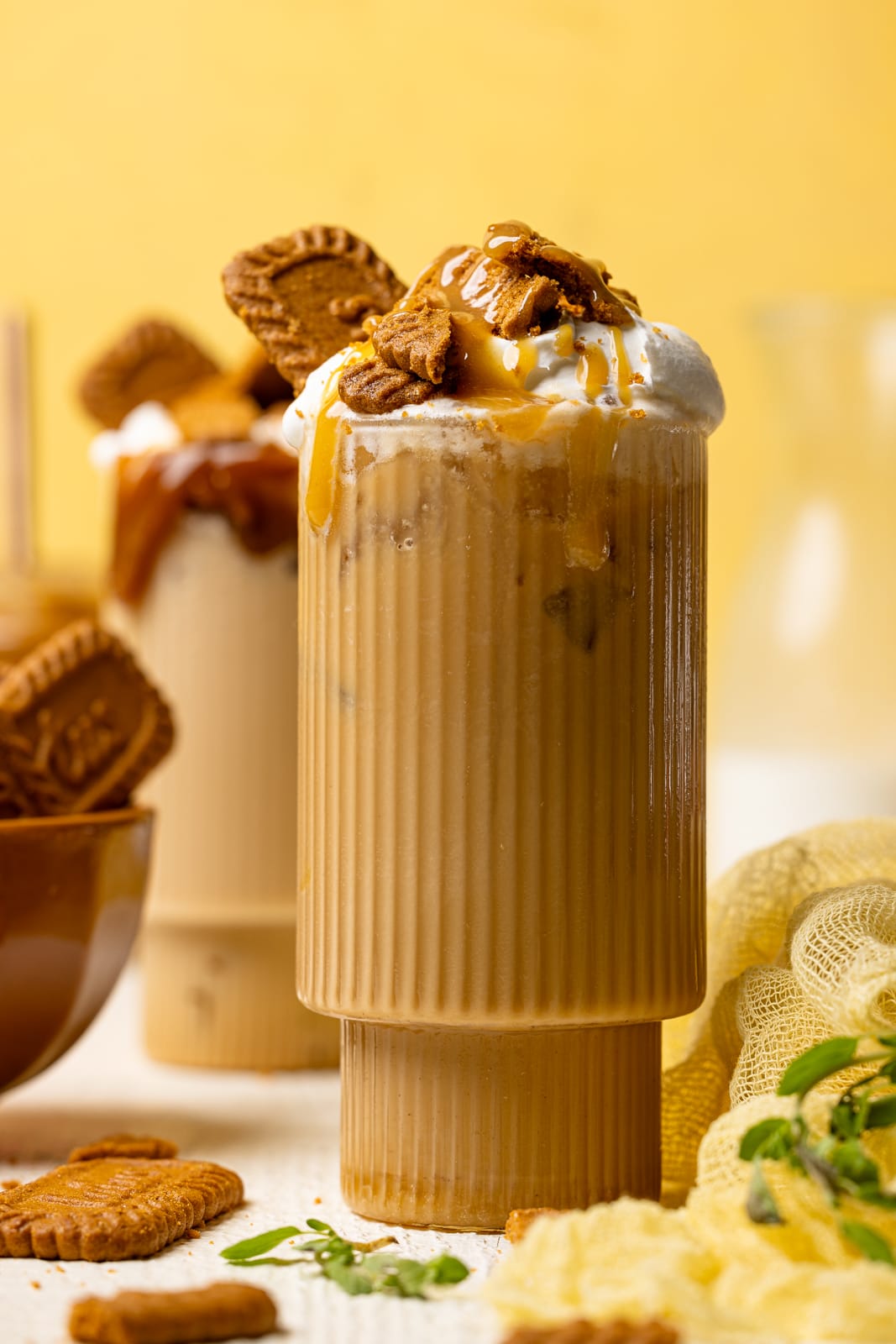 Latte in two tall glasses on a white table with biscoff cookies and a yellow background.