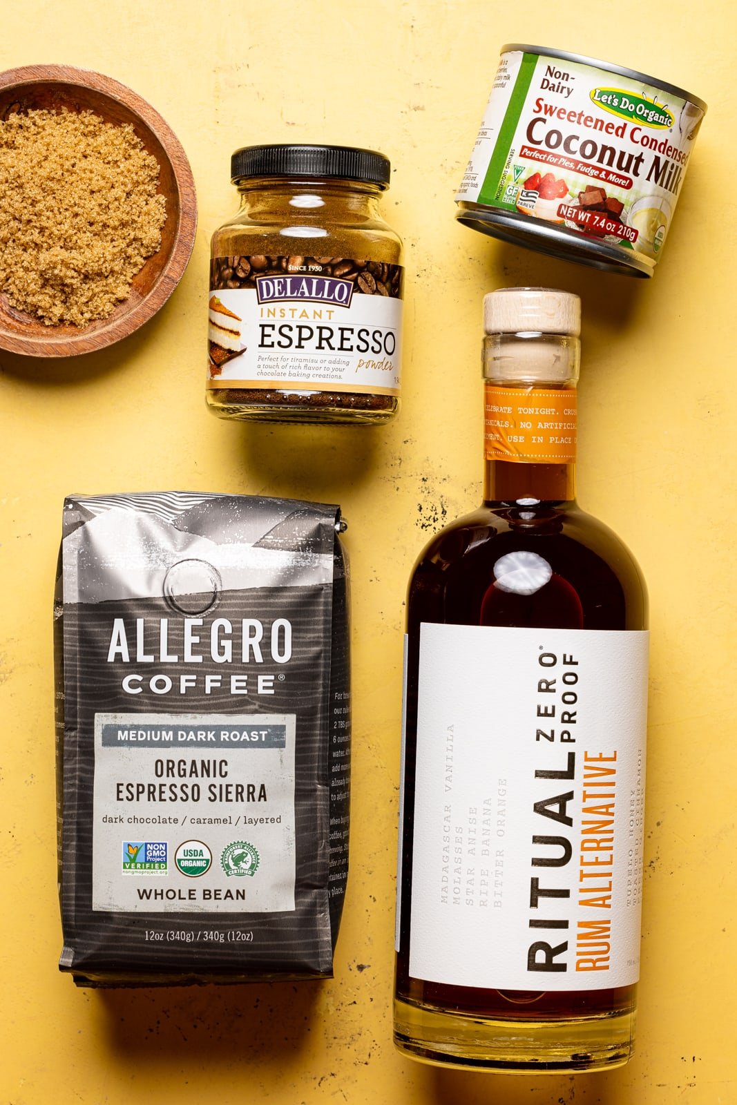Ingredients on a yellow table including brown sugar, Rum alternative, espresso coffee beans, dairy-free condensed milk, and espresso powder. 