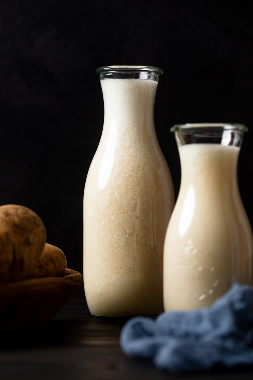 Two glass carafes of potato milk next to a bowl of potatoes