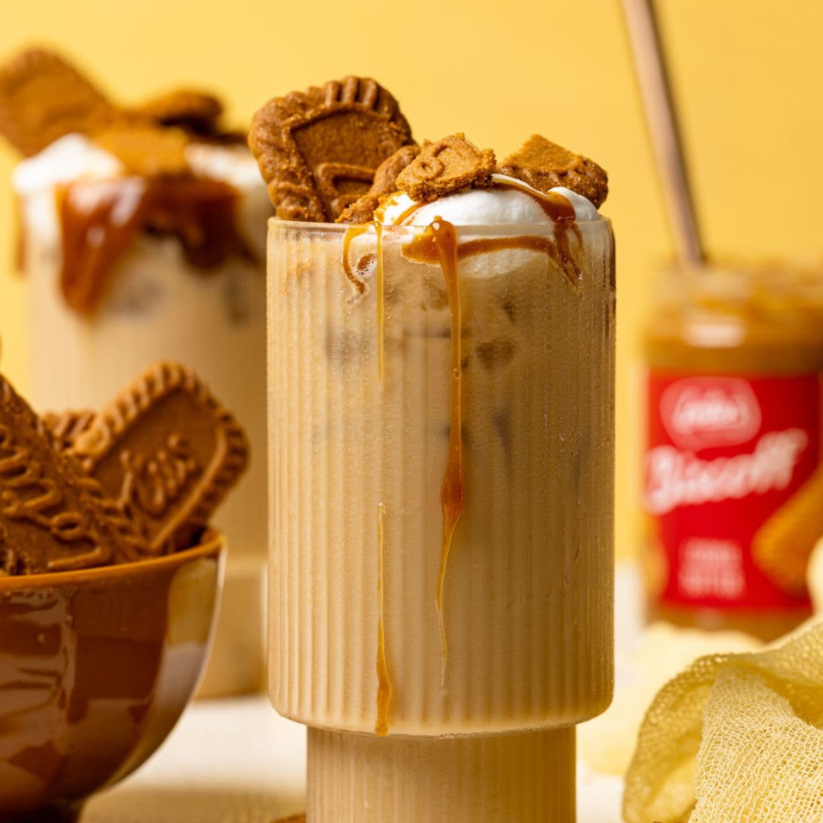 Two glasses with lattes, biscoff cookies, and cookie butter in the background on a white table and yellow background.