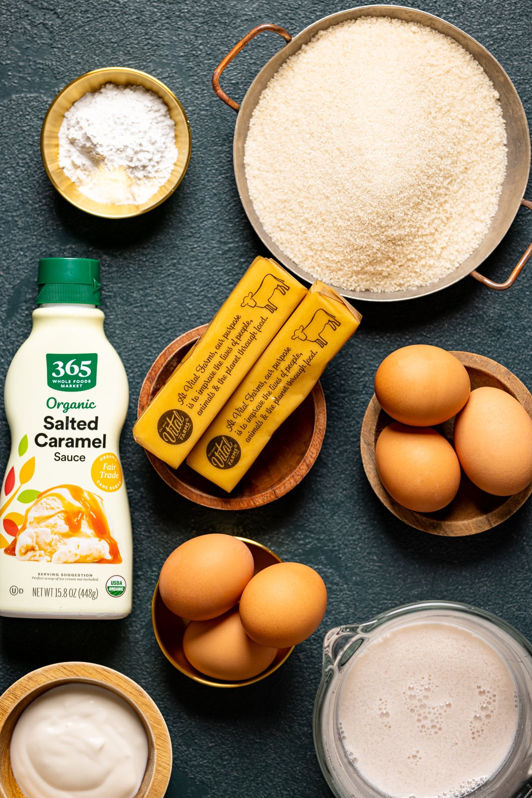 Ingredients on a dark green table including sugar, butter, eggs, salted caramel sauce, baking powder, and milk.