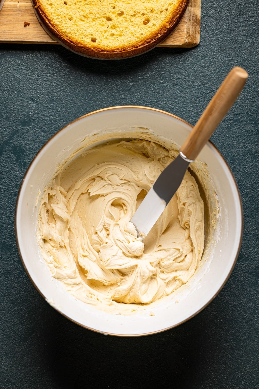 Buttercream in a white bowl with a spatula on a dark green table.