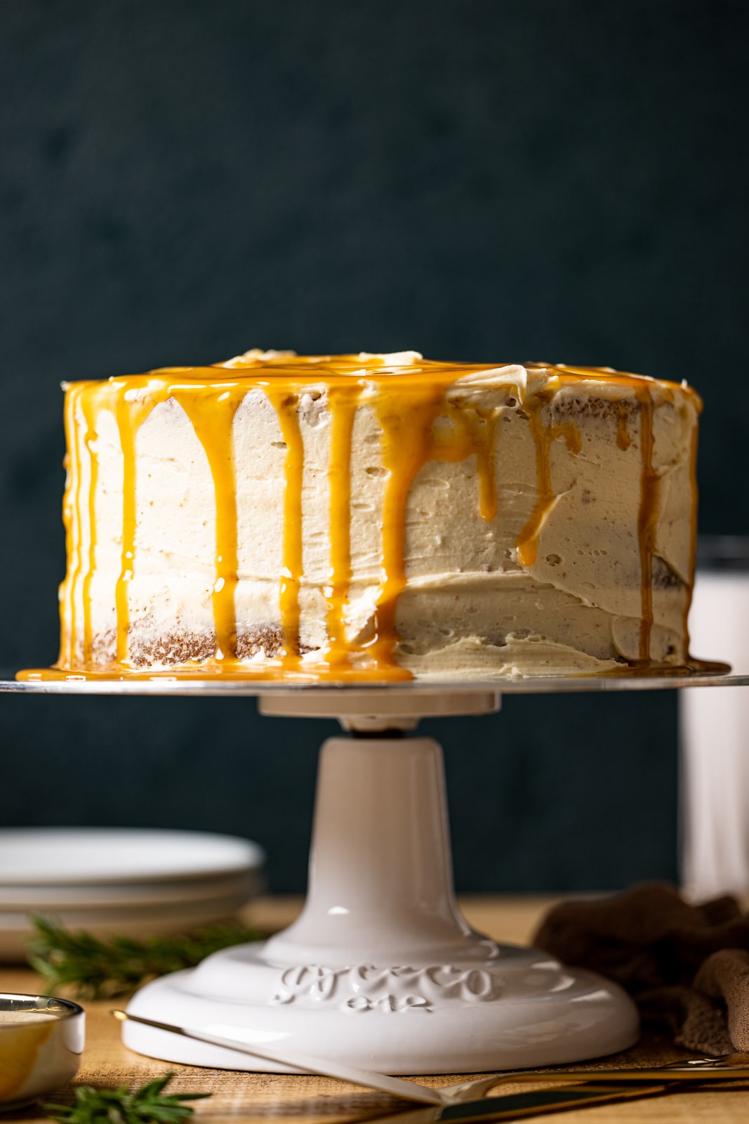Cake on a cake stand with drips of salted caramel on the tops and sides on a brown wood table with a dark green background.
