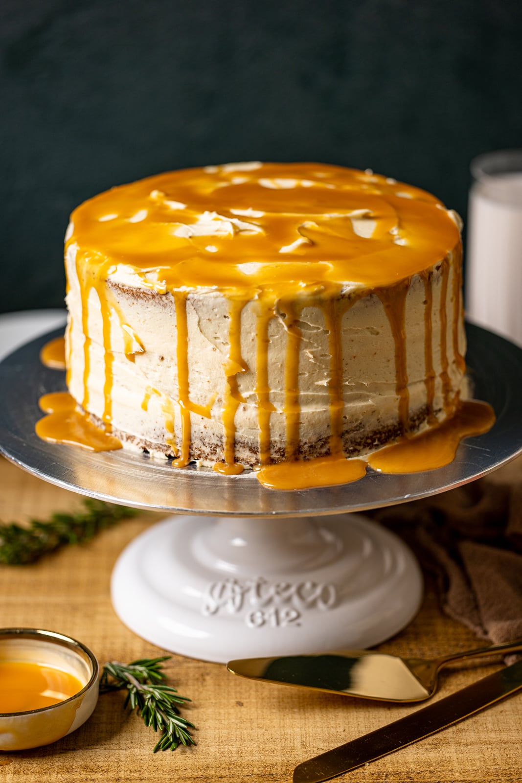 To view of fully frosted cake with drips of salted caramel on top on a cake stand on a brown table with a dark green background.