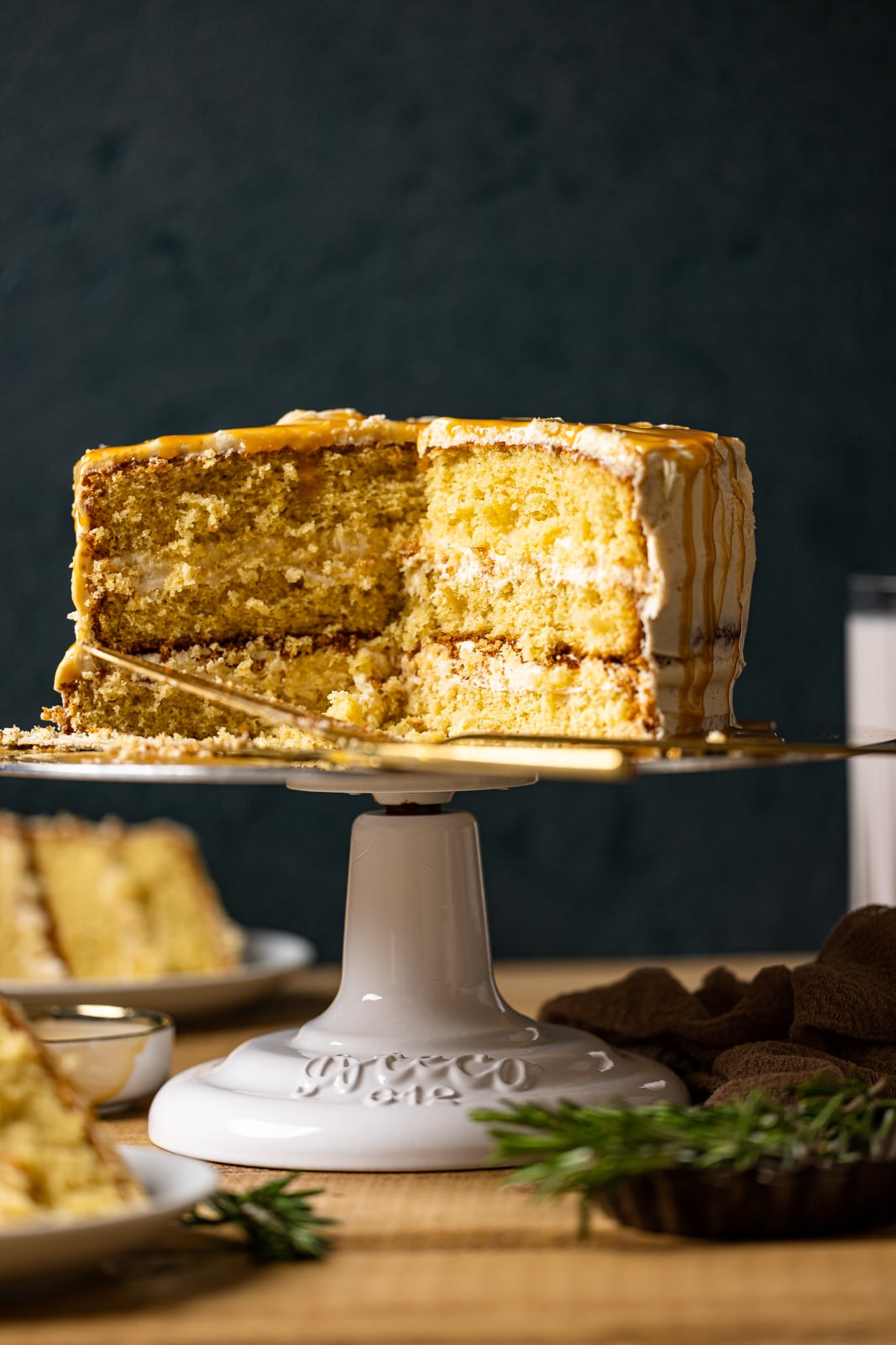 Whole caramel cake on a cake stand with slices surrounding it on a brown wood table and dark green background.