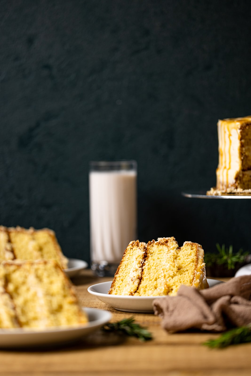 Slices of cake on white plates side by side with the cake stand in background along with a glass of milk on a brown wood table.