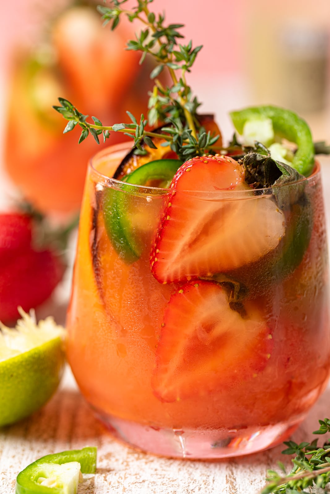Closeup of a Spicy Grapefruit Jalapeño \"Paloma\" Mocktail in a small glass filled with halved strawberries, jalapeno slices, and thyme