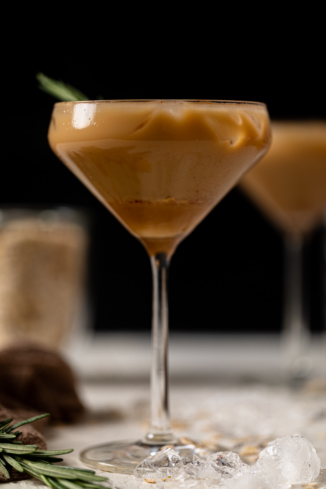 Caramel Oat Latte Mocktail in a long-stemmed glass near crushed ice and a sprig of rosemary