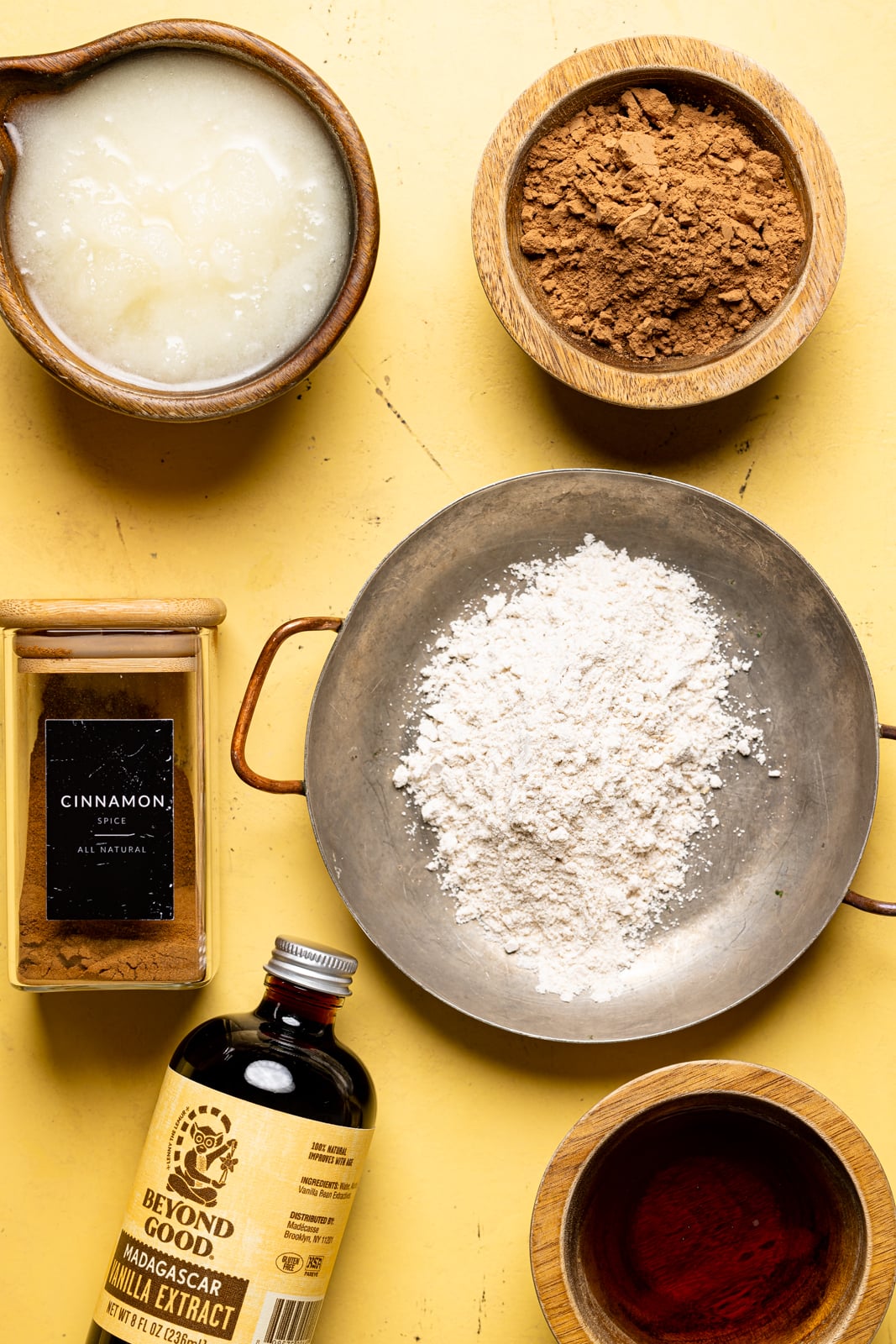 Ingredients on a yellow table including cocoa powder, oat milk powder, cinnamon, vanilla, maple syrup, and coconut oil.