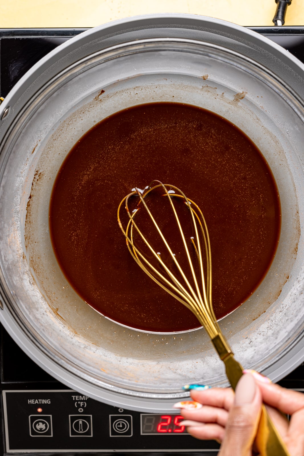 Melted chocolate in a large clear bowl with a gold whisk atop a skillet on the stovetop.
