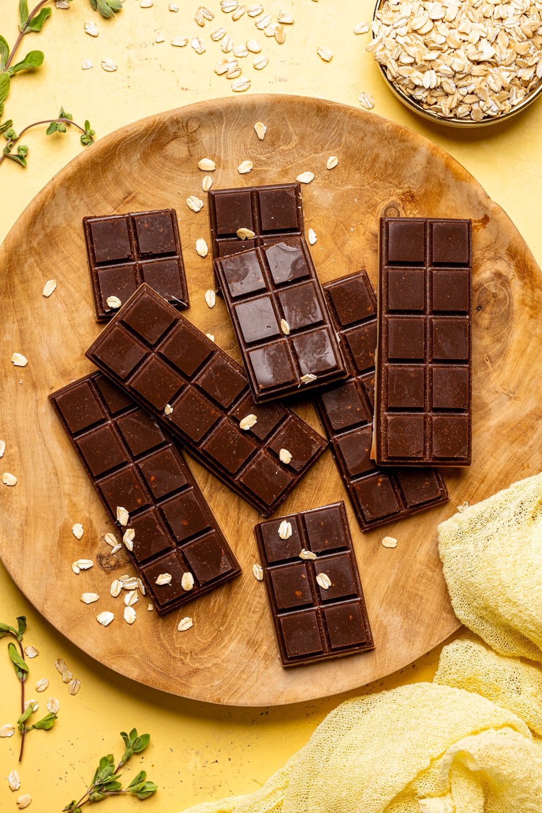 Chocolate bars on a brown wood plate with dry oats on a yellow table with a yellow napkin.